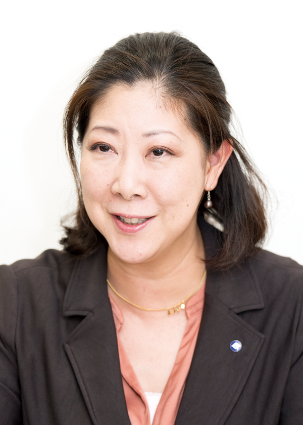 A professional portrait of a businesswoman with medium-length dark hair, wearing a dark blazer and a peach blouse, against a white background.