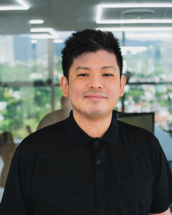 A portrait of a person in a black shirt standing in a modern office with a view of greenery and cityscape outside the windows.