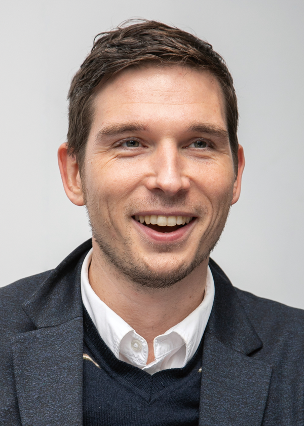 Portrait of a smiling man wearing business attire, including a dark blazer, white dress shirt, and sweater, against a plain background.