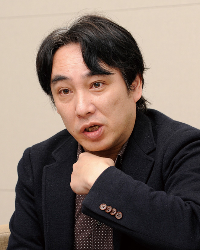 A person in a dark blazer is speaking and gesturing with their hand during an interview, shot in a neutral indoor setting.