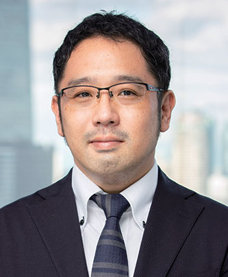 Portrait of a man in a suit and tie wearing glasses, photographed in front of a blurred office cityscape background. This image is for the Mori Building case study speaker profile.