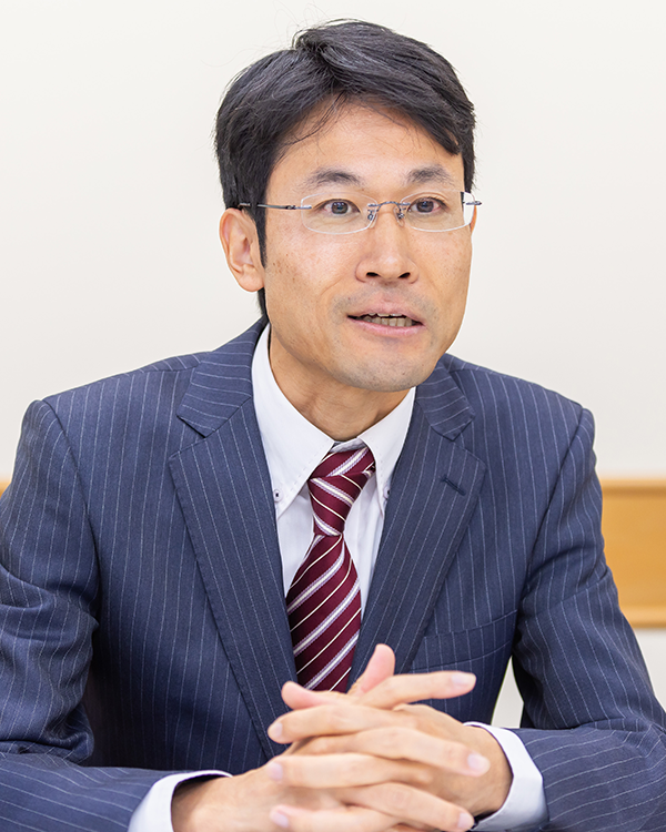A business professional in a suit and tie, seated and speaking during an interview. The image is associated with an NTT East case study for AWS Japan.