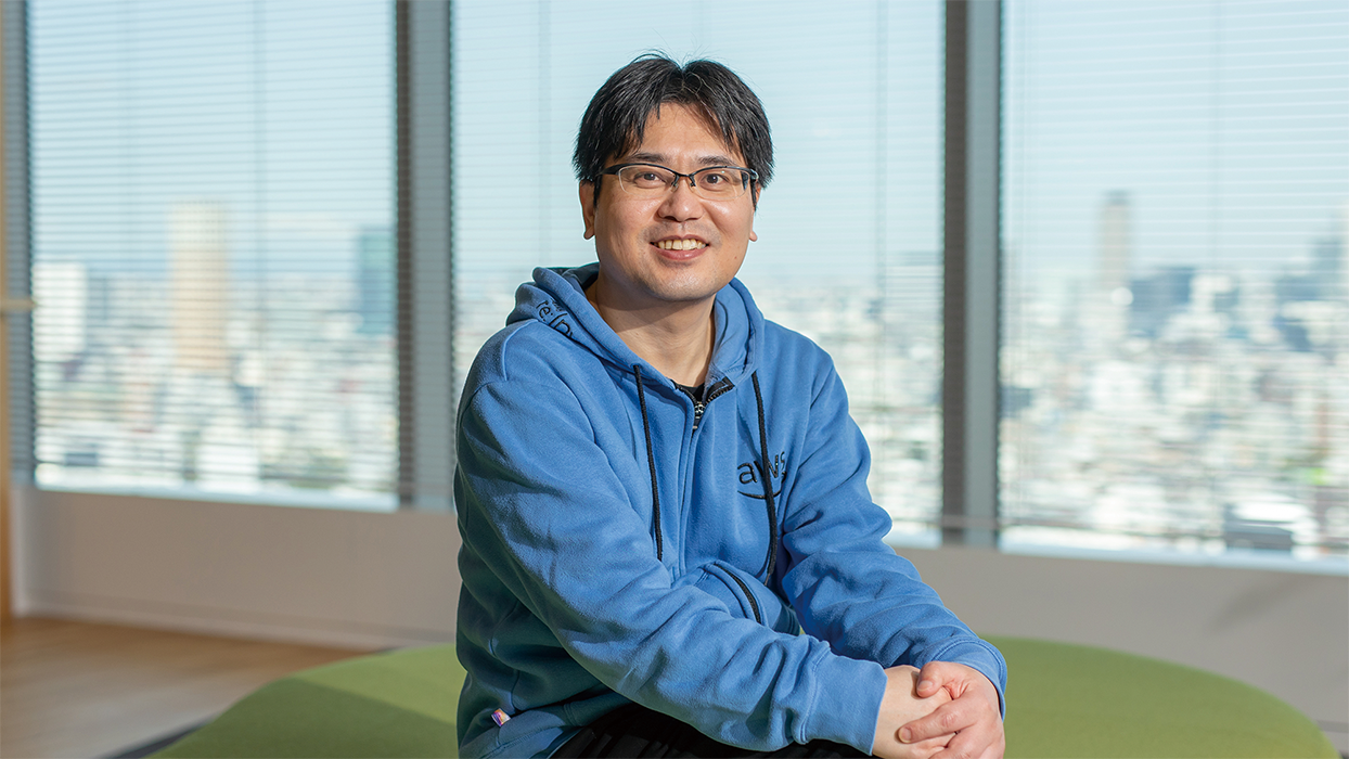 A man wearing a blue hoodie is smiling and sitting in a modern office setting with large windows and a cityscape blurred in the background.