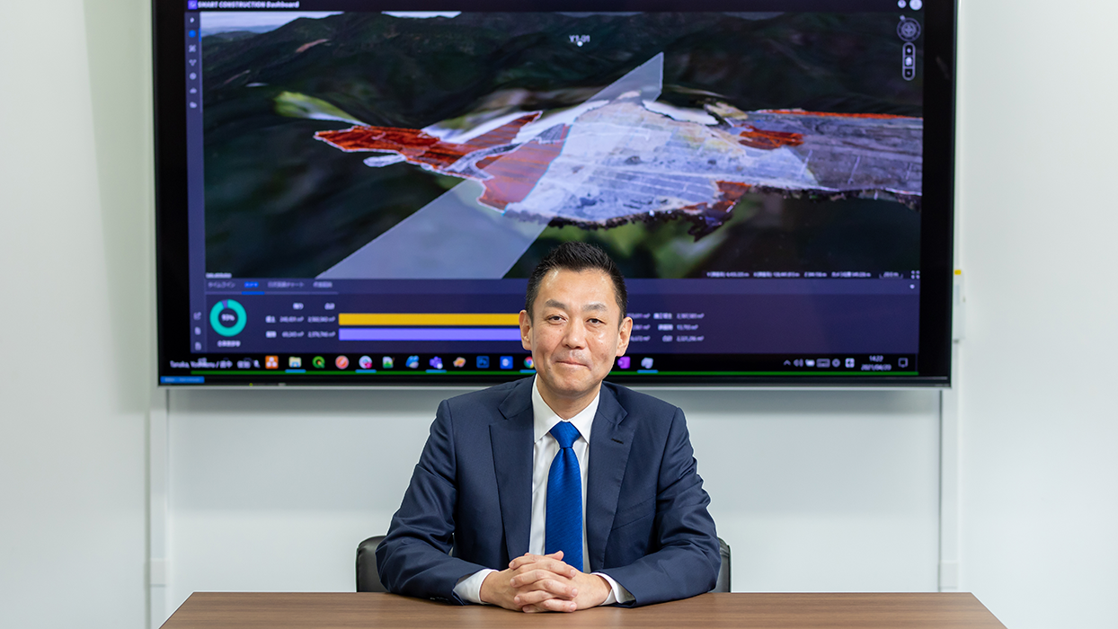 A businessman in a suit is sitting at a desk with hands folded, in front of a large digital dashboard screen displaying a 3D map and analytics, in an office environment. The digital dashboard in the background is labeled 'SMART CONSTRUCTION Dashboard' and is associated with Komatsu.
