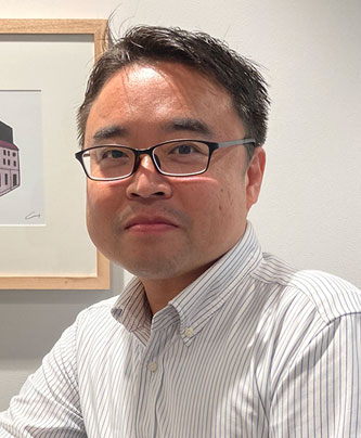 Portrait of a JPX case study speaker, Iwamura, wearing glasses and a white striped shirt, seated indoors with artwork in the background.