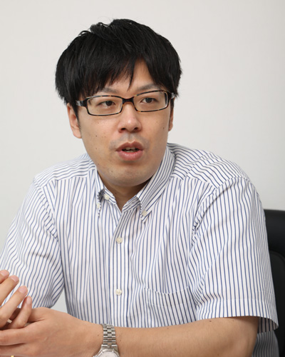 A man wearing a striped shirt and glasses is speaking during an interview, seated at a table.