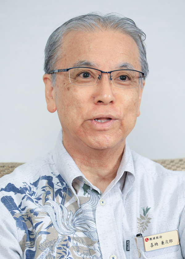 Portrait of a senior man wearing glasses and a patterned shirt, photographed indoors against a plain background. The man is facing forward and appears to be talking.