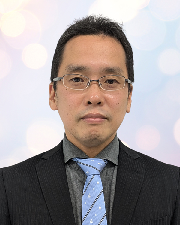 A person wearing a black pinstripe suit, gray shirt, and light blue tie with sailboat patterns, set against a soft, bokeh background.