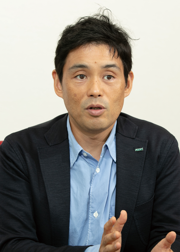 A portrait of a man in a suit jacket and light blue shirt, speaking during an interview. The setting appears to be professional and formal.