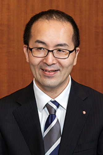 Portrait of Mr. Takahashi from Nihon Shokken, wearing a dark suit, white shirt, and striped tie, with glasses, posed against a wooden background.