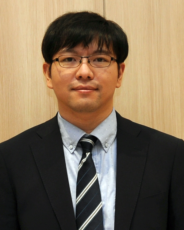 A man wearing glasses, a black suit, and a striped tie stands in front of a light wood background, posing for a professional headshot.