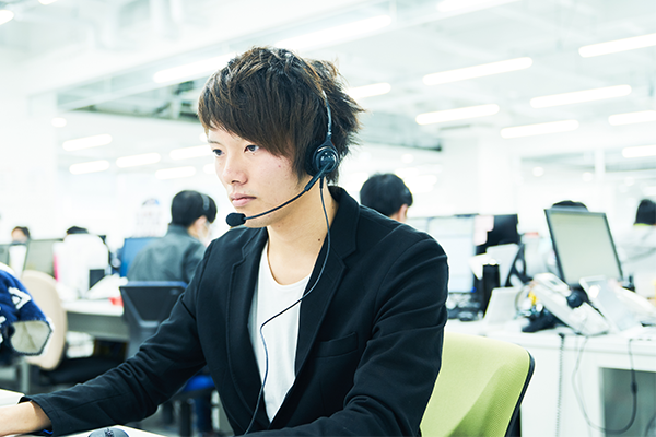 A call center agent wearing a headset works at a computer in a modern office environment. Other employees and computers are visible in the background.