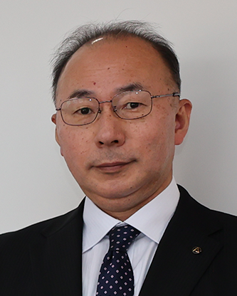 A professional portrait of a man wearing a suit, tie, and glasses, standing against a plain background.