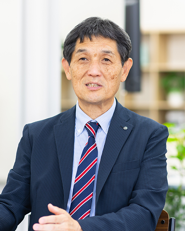 A professional portrait of a man in a suit and tie being interviewed, set in an office environment. This image is associated with Yamanashi Chuo Bank and features Tanaka for a solution case study interview.