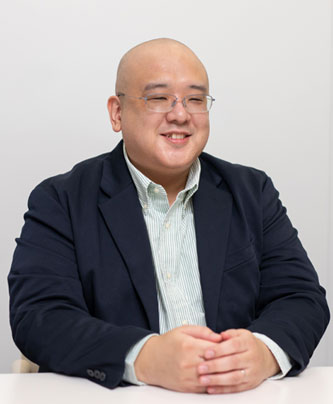 Portrait of an East Asian man wearing glasses and a dark suit jacket, seated and smiling, against a plain background. Professional speaker photo for Sony Interactive Entertainment.