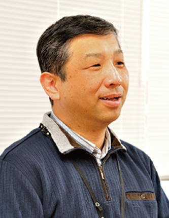 A person wearing a navy blue sweater with a collared shirt underneath, sitting indoors near a window with blinds.