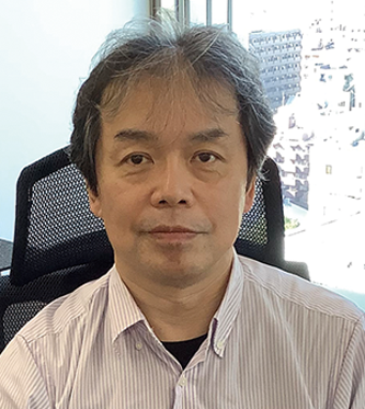 Portrait of a person sitting in an office setting, associated with Transcosmos, featuring a neutral expression and city buildings visible in the background.