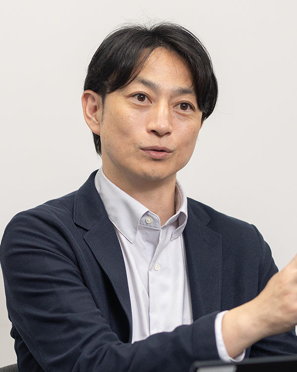 A business portrait of a man with dark hair wearing a white collared shirt and dark blazer, gesturing during a discussion against a plain background.