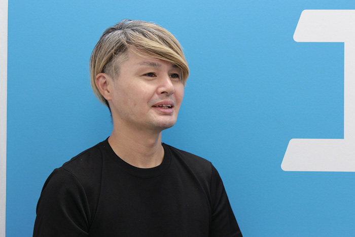 A person with light blonde hair wearing a black shirt, posing in front of a blue background with white graphic elements.