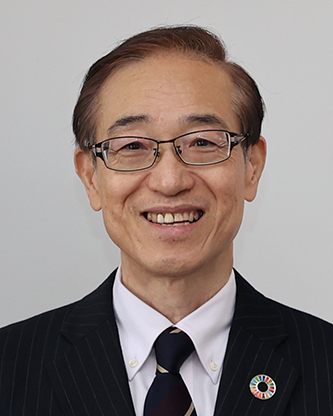Portrait of a smiling man in a suit and tie, wearing glasses and a pin, against a plain background.