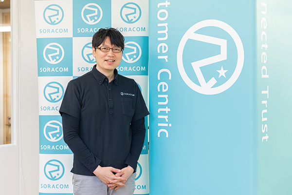 Portrait of Mr. Katayama, a Soracom customer, standing and smiling in front of a Soracom logo backdrop and a banner with company branding.