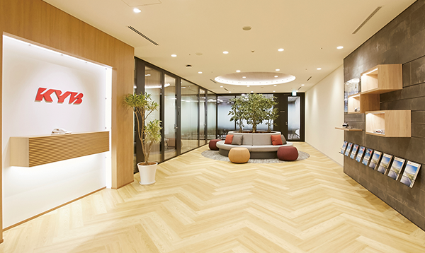 Modern office lobby with light wood flooring, a reception desk with the KYB logo, seating area with plants, and shelves displaying brochures.