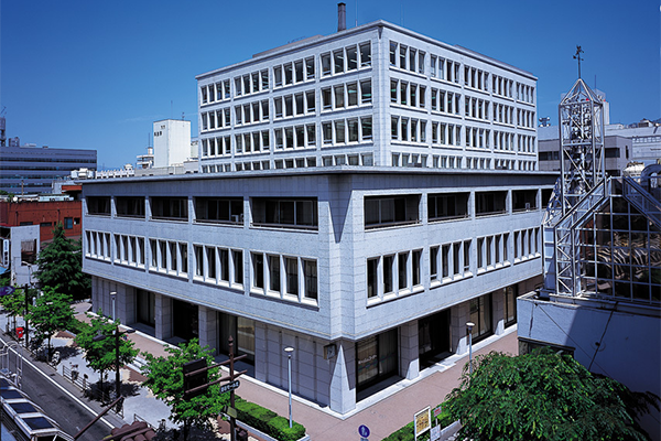 A modern, multi-story office building with white and gray exterior, surrounded by trees and urban structures under a clear blue sky.