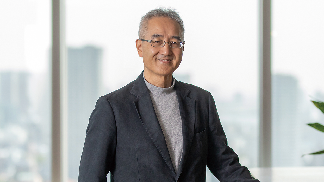 A businessman wearing glasses and a dark blazer, standing in a modern office with large windows and a blurred cityscape in the background.