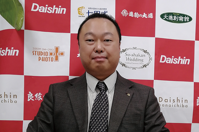 A business portrait of a man in a suit sitting in front of a step-and-repeat background featuring multiple Daishin and partner logos. Suitable for use in a case study or corporate publication.