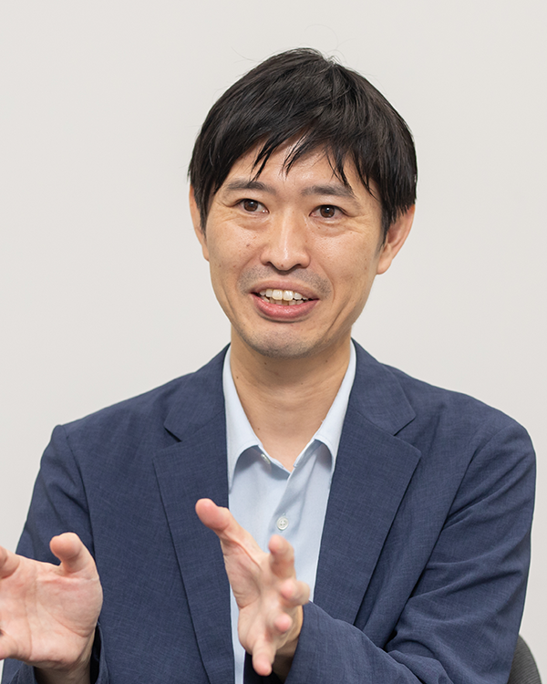 A professional portrait of a man in a blue suit and light blue shirt, engaging in conversation or an interview, with a neutral background.