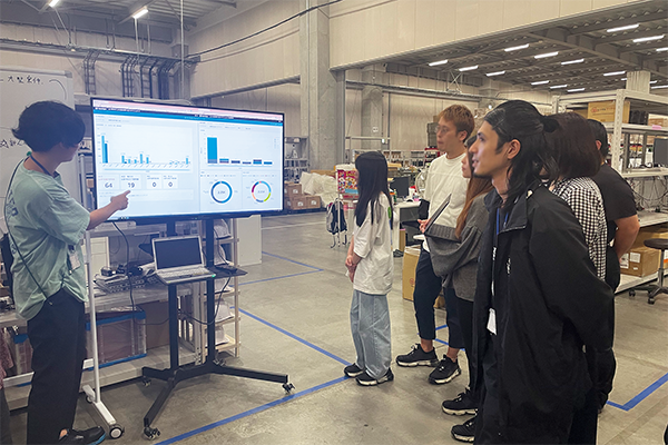 A group of people in a warehouse setting observing a presentation on a large screen showing data analytics dashboards. The presenter is explaining various charts and graphs to the audience.