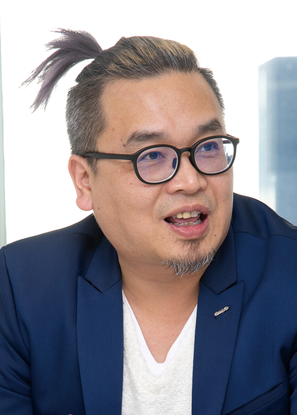 A man with glasses, a blue blazer, and a ponytail is talking or engaged in conversation.