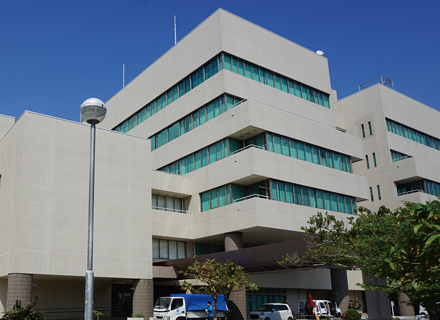 A modern office building with large windows and beige exterior is shown under a clear blue sky during daytime. The setting includes a lamp post, several trees, and vehicles parked in front of the building.
