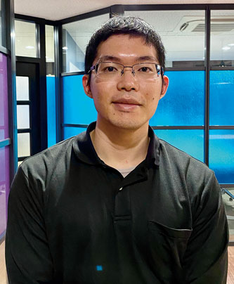 A professional office portrait of a man with glasses wearing a black shirt, standing in an office setting with glass walls and blue and purple panels.