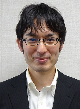 A person wearing a black suit jacket and white collared shirt against a light gray background.