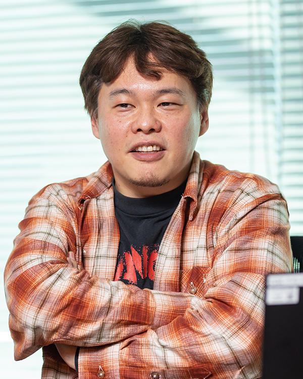 Portrait photo of a Bandai Namco case study speaker in a casual shirt with arms crossed, seated indoors with window blinds in the background.