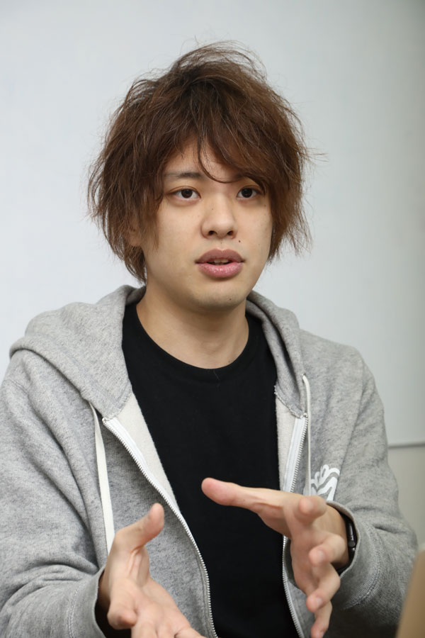 A young person in casual clothing, including a gray hoodie and black shirt, is sitting and gesturing with their hands while speaking during an interview, with a neutral background.