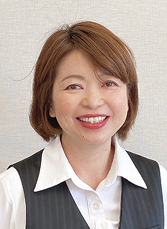 A professional portrait of a smiling woman with short brown hair, dressed in a white shirt and dark vest, standing in front of a neutral background.