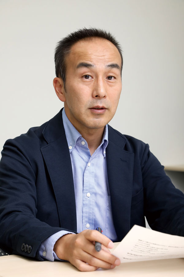 A portrait of a business professional seated at a desk, holding a pen and a piece of paper, dressed in a blue shirt and dark blazer.