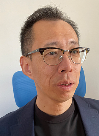 Portrait of a man with glasses sitting in front of a blue chair, looking slightly to the side. He is wearing a dark blazer and black shirt.