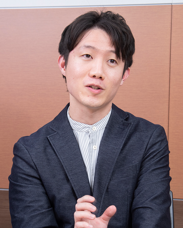 A person wearing a dark blazer and striped shirt, gesturing with one hand, against a wooden panel background.