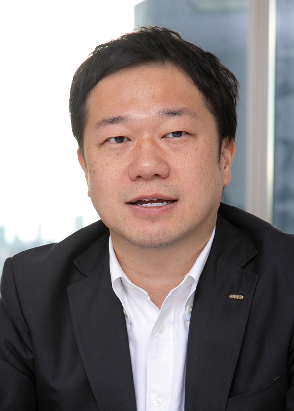 A portrait of a businessman in a suit, seated in an office setting with large windows and cityscape in the background.