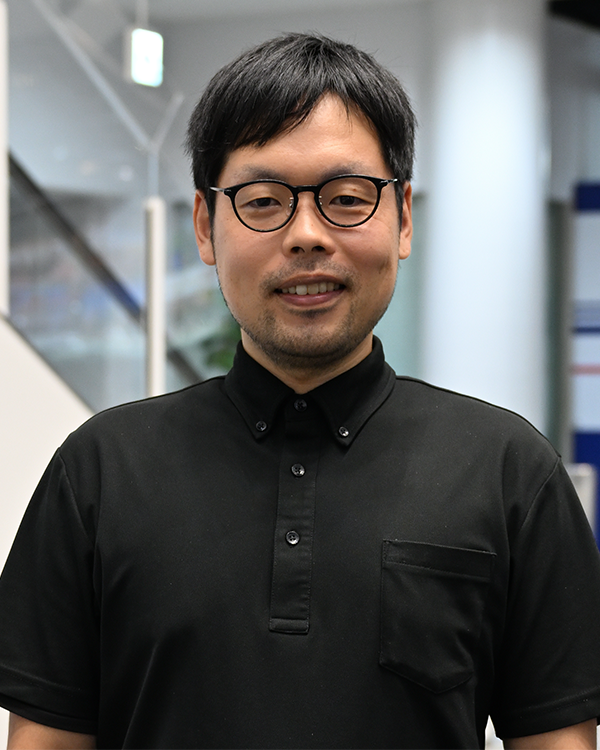 A person wearing a black buttoned shirt, standing indoors with a blurred background featuring stairs and glass panels.
