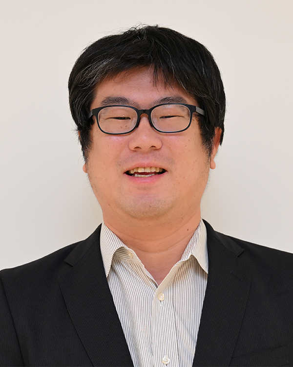 A person wearing a black suit jacket and a light striped collared shirt against a plain background.