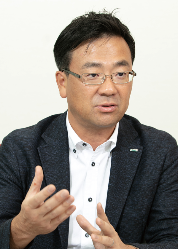 A man in a suit and glasses speaking during an interview, gesturing with his hands. White background. (600x840 image)