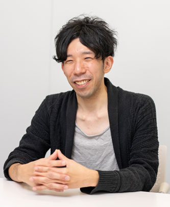 A man seated at a white table, smiling, dressed casually in a dark sweater and gray shirt, in a modern, neutral-colored setting. This image is used for a solution case study for Sony Interactive Entertainment on the AWS Japan website.