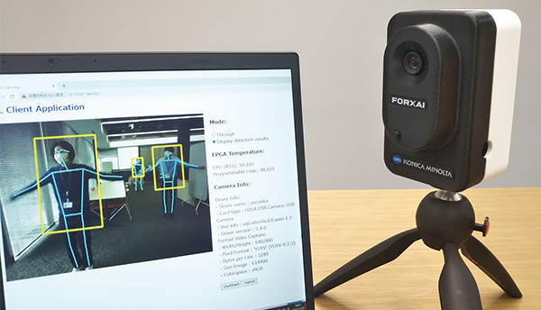 A Konica Minolta FORXAI AI camera is positioned on a tripod beside a laptop displaying real-time detection results, illustrating usage in an AWS case study.