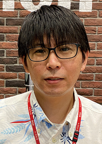 A person wearing glasses and a floral shirt with a red lanyard, standing in front of a brick wall. This image is used for an Orion Beer solution case study on the AWS Japan website.