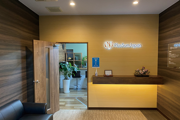 "Office reception area with a wooden desk, PlusSum Japan logo on the wall, and an open door revealing a room with plants and shelves."