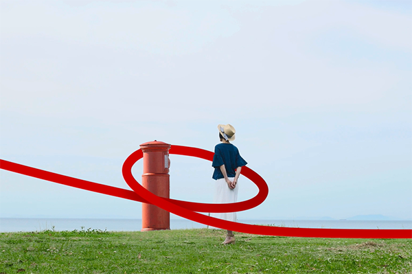 A woman in a hat and blue top stands on grass near a red postbox, with a swirling red ribbon encircling both against a backdrop of sky and sea.